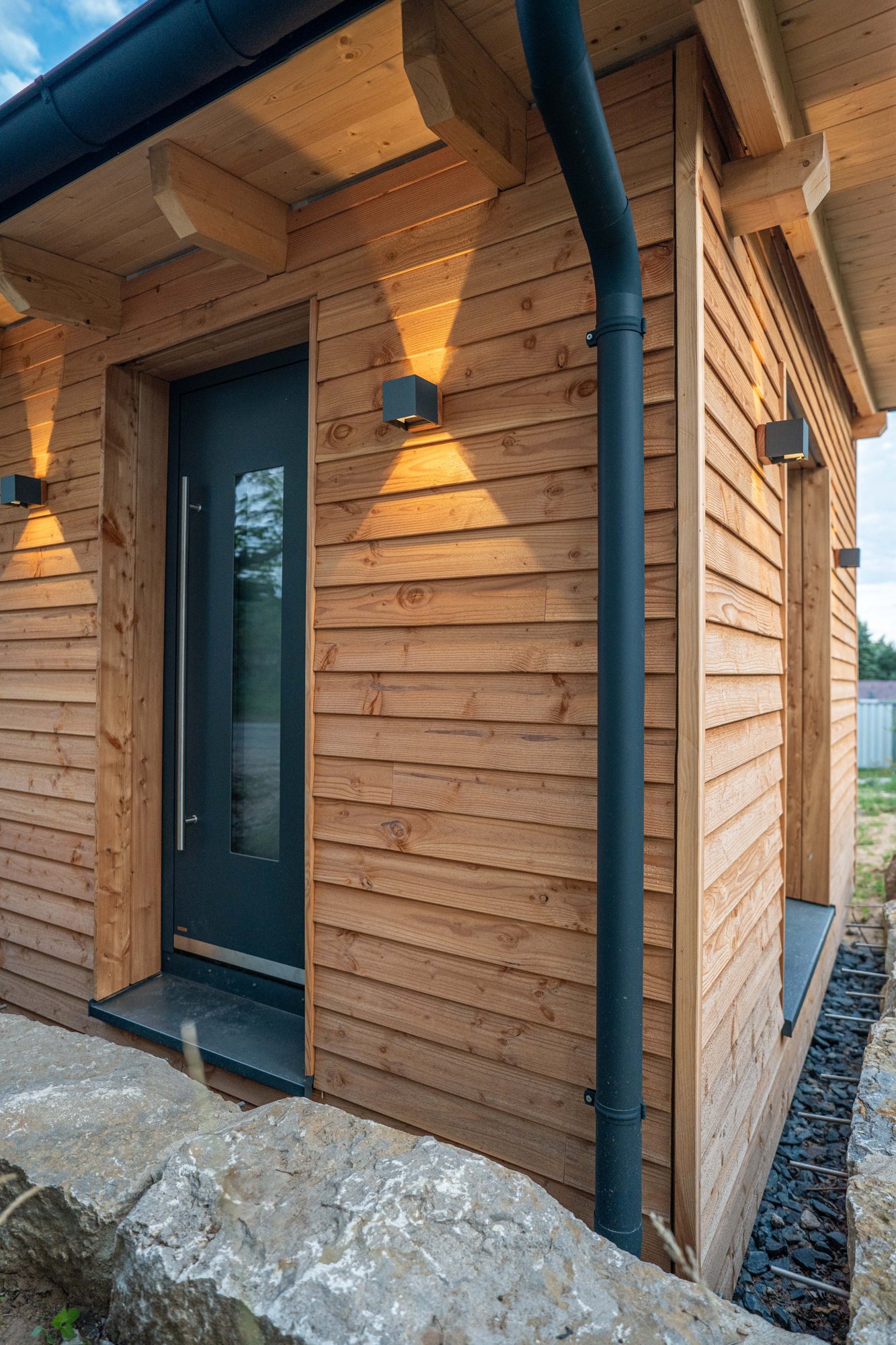 Eingang eines Holzhauses: Lärchenfassade mit Stülpschalung, anthrazitfarbene Haustür und warm leuchtende Wandleuchten